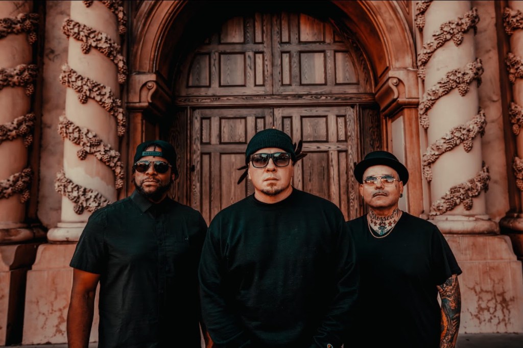 Three members of the band P.O.D. posed in front of a decorative building, wearing sunglasses and black clothing.